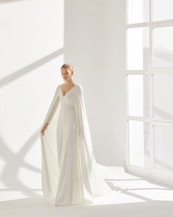 Préndate de Rima, un vestido de dos piezas formado por un traje y una capa de crepé fluida. Una boda eclesiástica en otoño es el marco ideal para lucir este modelo de línea A y escote de pico. Su sencilla manga larga compensa a la perfección la espalda descubierta y la cintura bordada. Tiendas de vestidos de Novia en Toledo, El Clavel