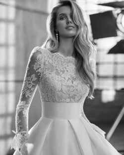 Modelo Walta - Vestido de novia largo, de corte evasé y estilo clásico, confeccionado en encaje rebrodé y mikado Magnolia. Presenta un escote barco, una espalda en V y manga larga. Se completa con una falda de maxi volumen con fajín, bolsillos y pliegues. Un modelo Aire Atelier de color natural ideal para una boda religiosa en invierno. Vestidos de novia El Clavel Toledo