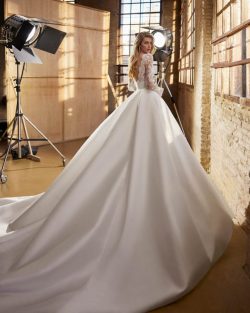 Modelo Walta - Vestido de novia largo, de corte evasé y estilo clásico, confeccionado en encaje rebrodé y mikado Magnolia. Presenta un escote barco, una espalda en V y manga larga. Se completa con una falda de maxi volumen con fajín, bolsillos y pliegues. Un modelo Aire Atelier de color natural ideal para una boda religiosa en invierno. Vestidos de novia El Clavel Toledo