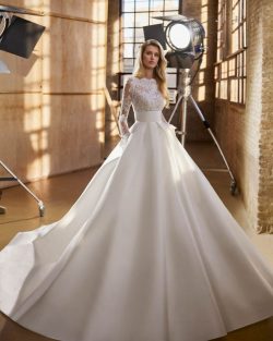 Modelo Walta - Vestido de novia largo, de corte evasé y estilo clásico, confeccionado en encaje rebrodé y mikado Magnolia. Presenta un escote barco, una espalda en V y manga larga. Se completa con una falda de maxi volumen con fajín, bolsillos y pliegues. Un modelo Aire Atelier de color natural ideal para una boda religiosa en invierno. Vestidos de novia El Clavel Toledo