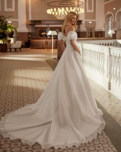 Colección vestidos de novia 2025 en El Clavel Toledo Vestido de novia largo, con silueta en línea A y estilo romántico, confeccionado en garza. Disfruta de un escote palabra de honor y manga con volantes extraíble. Un exclusivo modelo Aire Barcelona