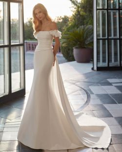Modelo Vincet Vestidos de novia largos de corte sirena y estilo moderno, elaborado con mikado. Presenta un escote que deja los hombros al descubierto, tirantes caídos, un lazo de adorno en la espalda y una sobrefalda extraíble. Un exclusivo conjunto que encontrarás en El Clavel Toledo