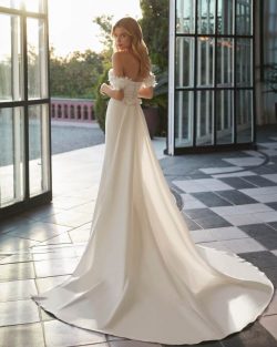 Modelo Vincet Vestidos de novia largos de corte sirena y estilo moderno, elaborado con mikado. Presenta un escote que deja los hombros al descubierto, tirantes caídos, un lazo de adorno en la espalda y una sobrefalda extraíble. Un exclusivo conjunto que encontrarás en El Clavel Toledo