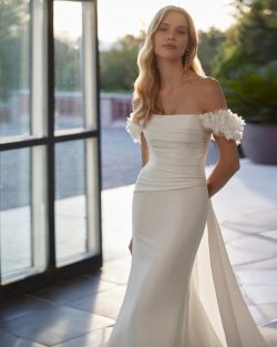 Modelo Vincet Vestidos de novia largos de corte sirena y estilo moderno, elaborado con mikado. Presenta un escote que deja los hombros al descubierto, tirantes caídos, un lazo de adorno en la espalda y una sobrefalda extraíble. Un exclusivo conjunto que encontrarás en El Clavel Toledo